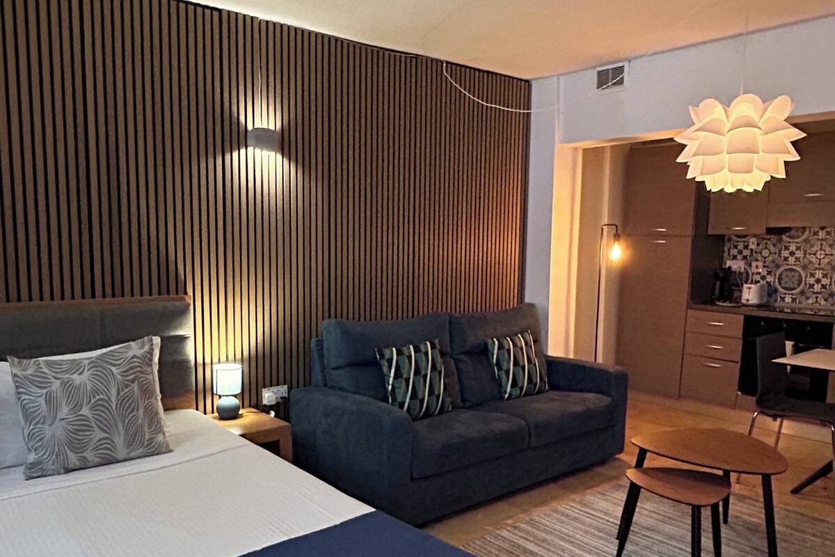 A modern hotel room with a large bed, a sofa, a dining table, and a kitchen area.