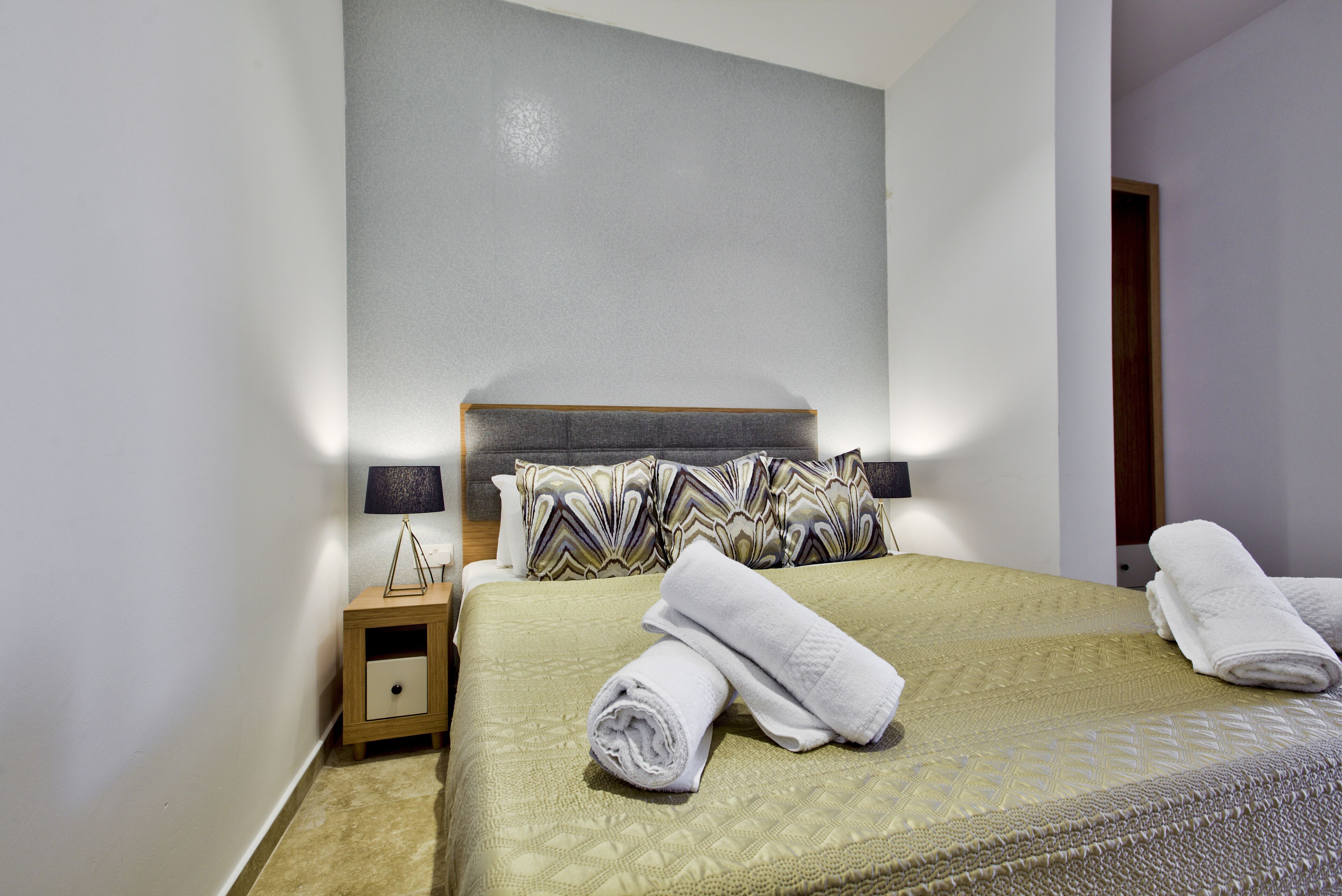 A neatly made bed with a green quilt, two pillows, and rolled white towels on a wooden nightstand.