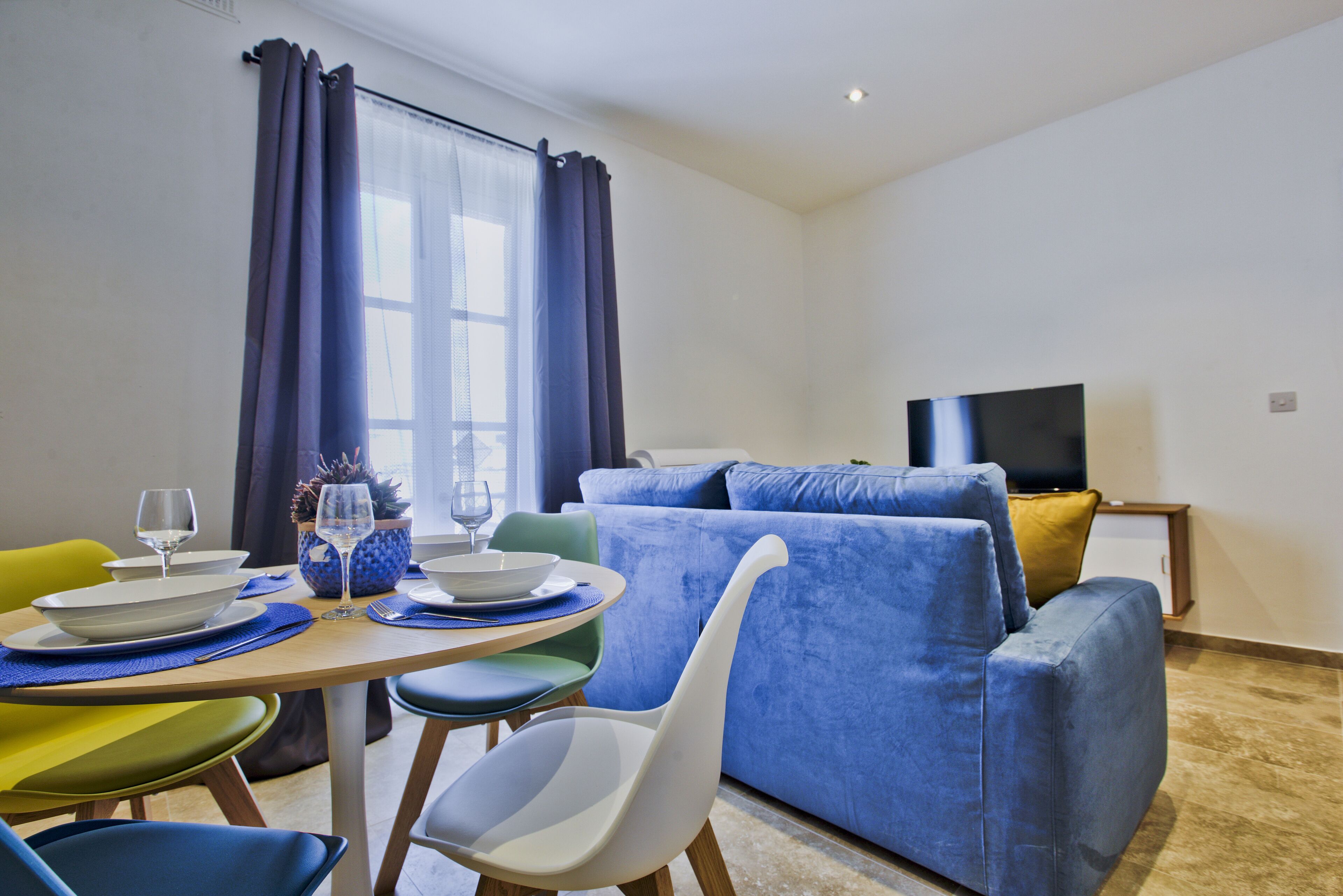 A modern living room with a dining area, featuring a blue sofa, a flat-screen TV, and a dining table set with plates, glasses, and cutlery.