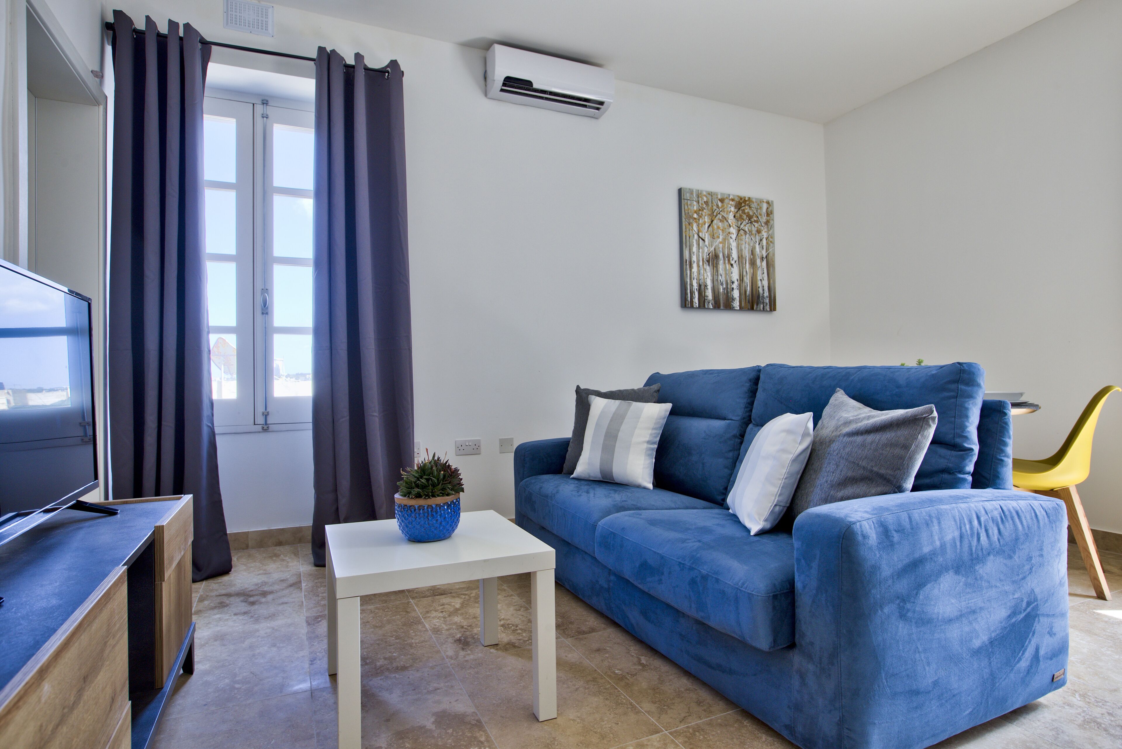 Un salon avec un canapé bleu, une table basse blanche, un téléviseur posé sur un meuble en bois, une plante en pot et un tableau accroché au mur.