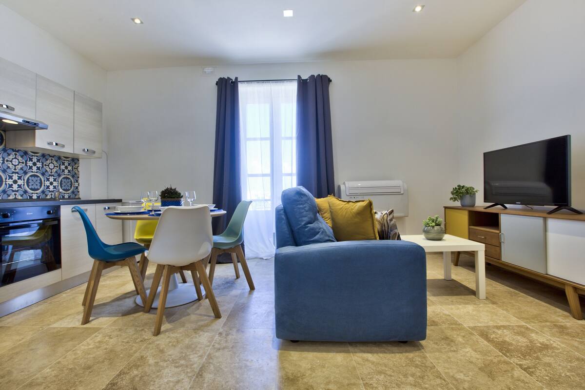A modern living room with a blue sofa, a dining table with chairs, a kitchen area with a stove, and a television.