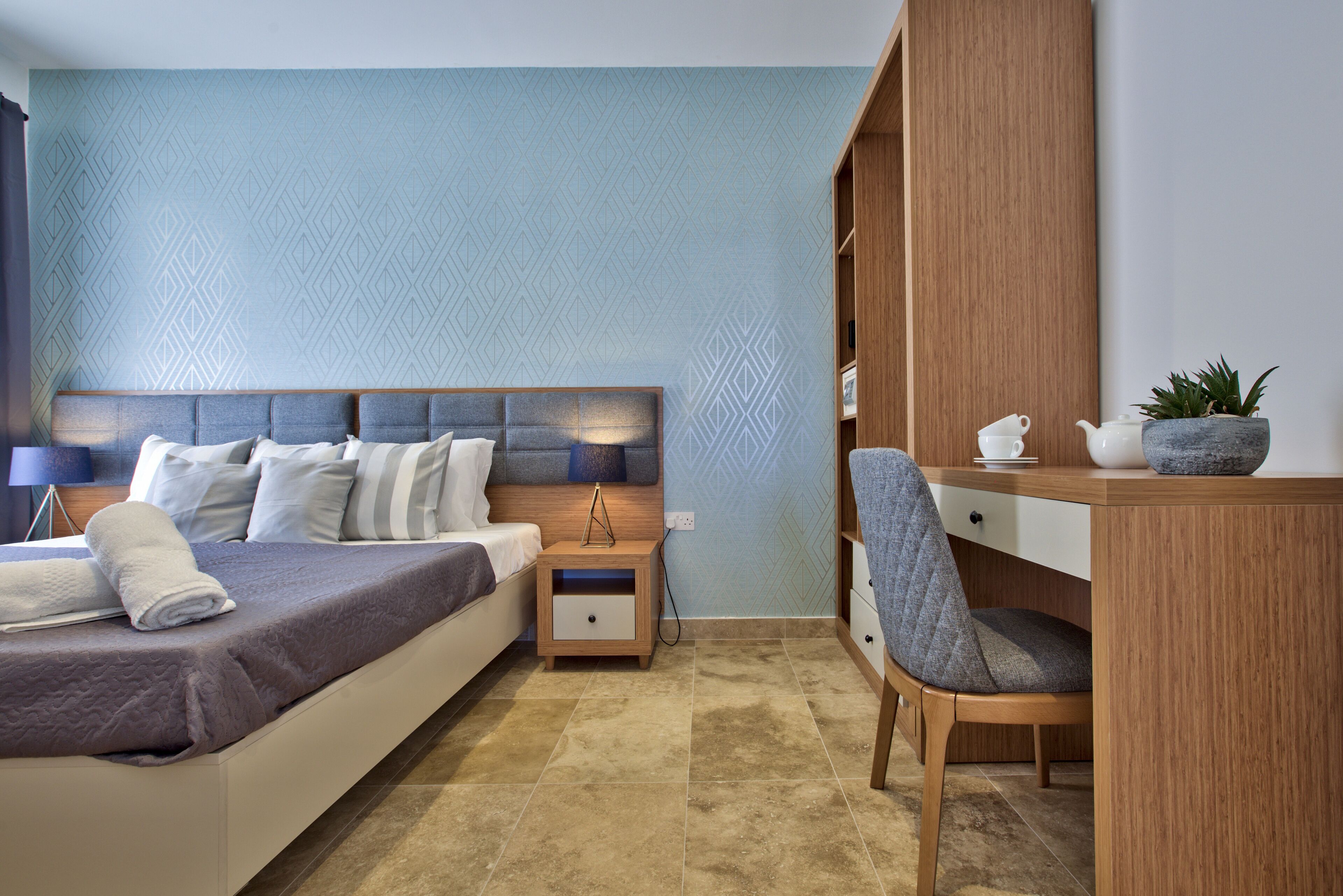 A modern bedroom with a bed, bedside table, chair, and a desk with a plant.