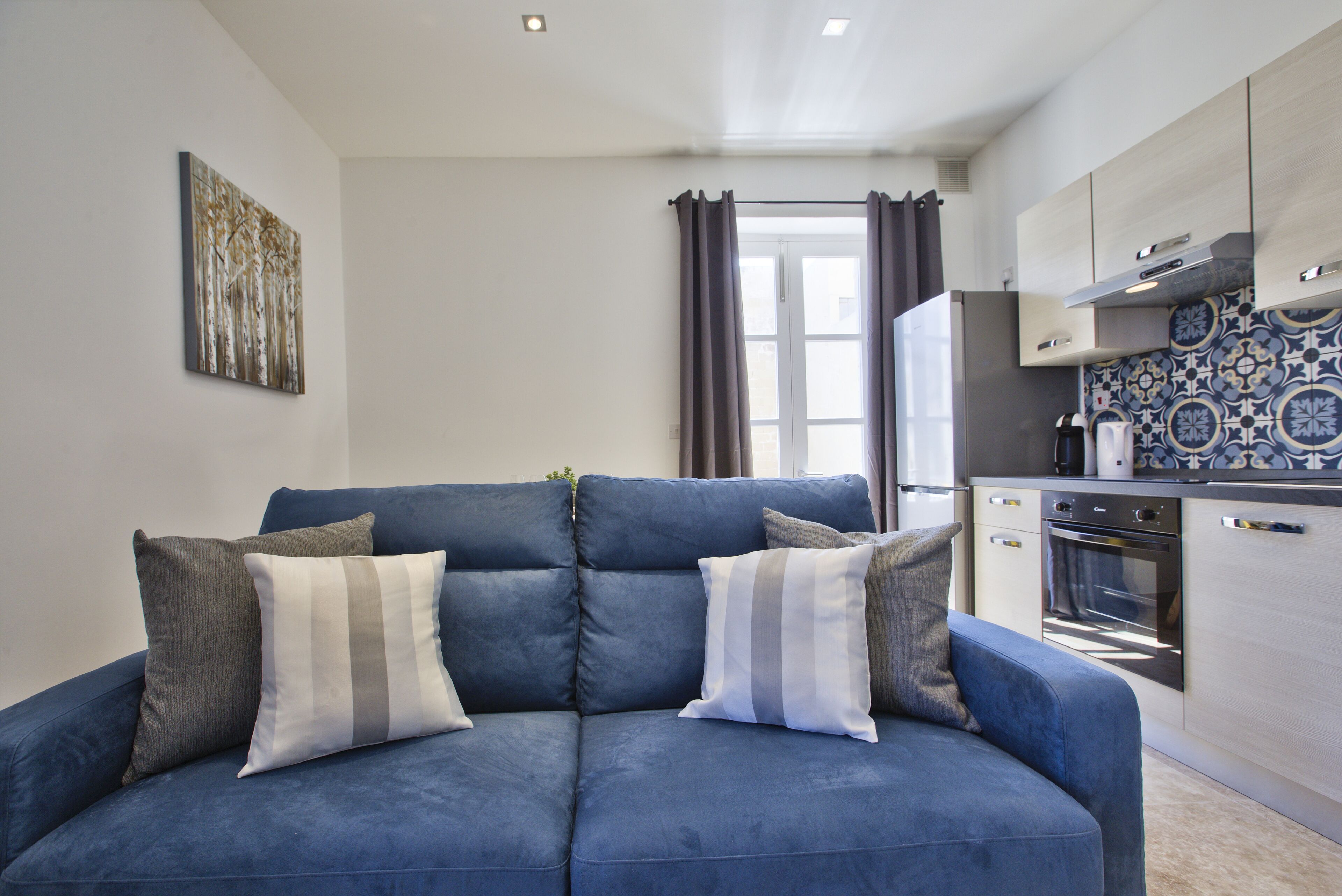 Un salón moderno con un sofá azul, una cocina con armarios de madera clara y una ventana con cortinas.