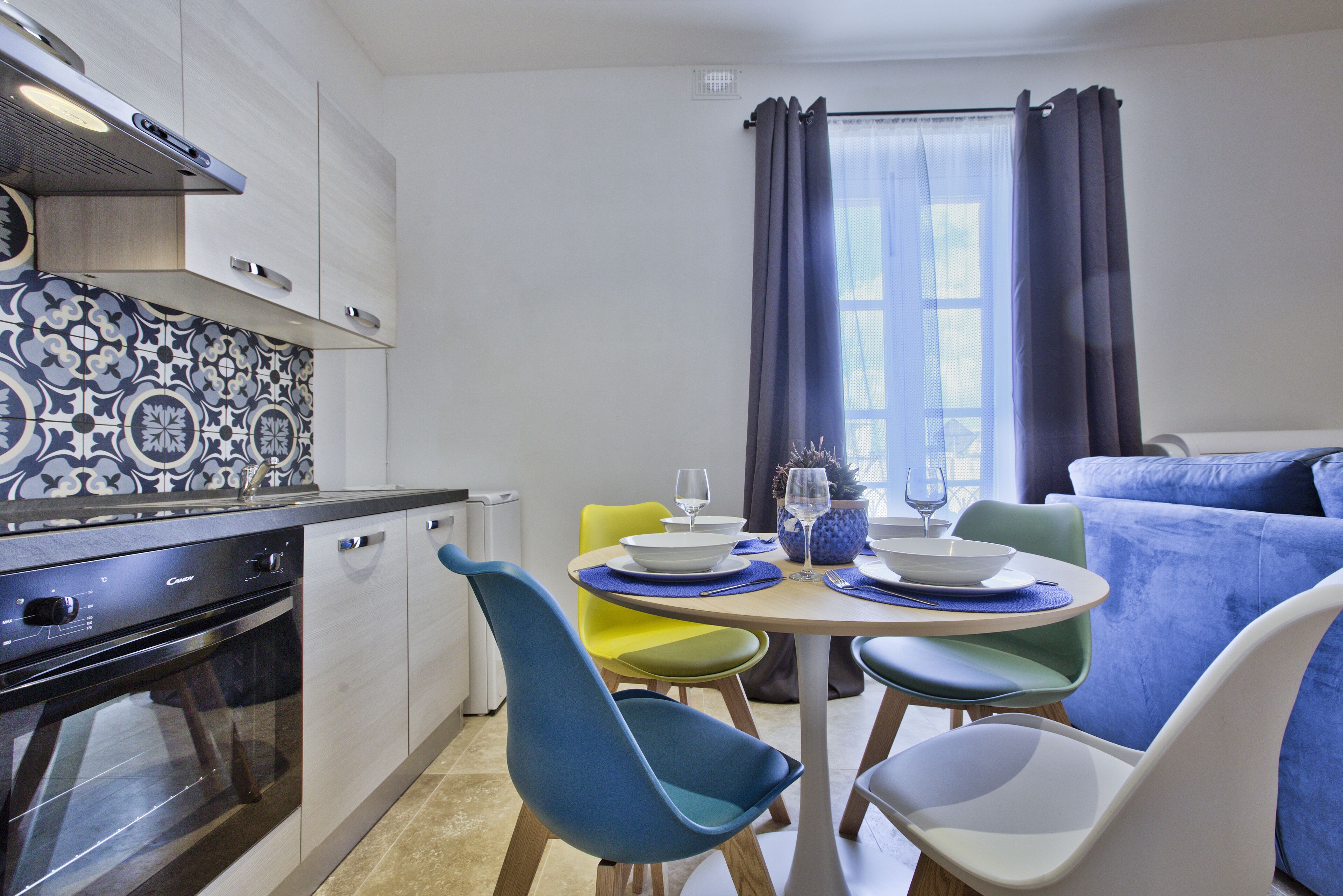 Una cocina moderna con horno empotrado, un comedor con sillas coloridas y un amplio ventanal con cortinas.