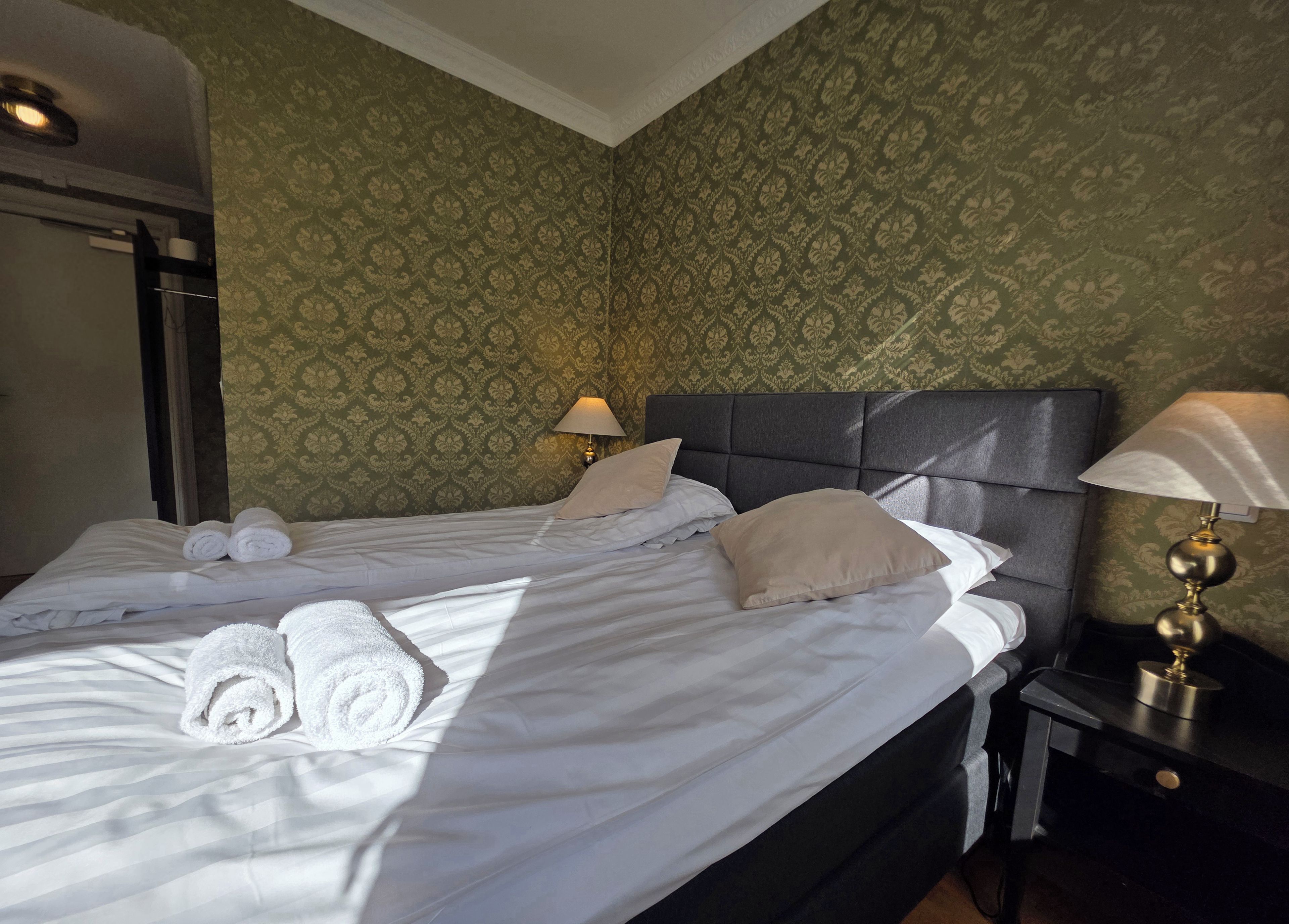 A neatly made bed with rolled towels, two bedside lamps, and a patterned wallpaper.