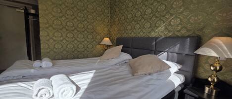 A neatly made bed with rolled towels, two bedside lamps, and a patterned wallpaper.