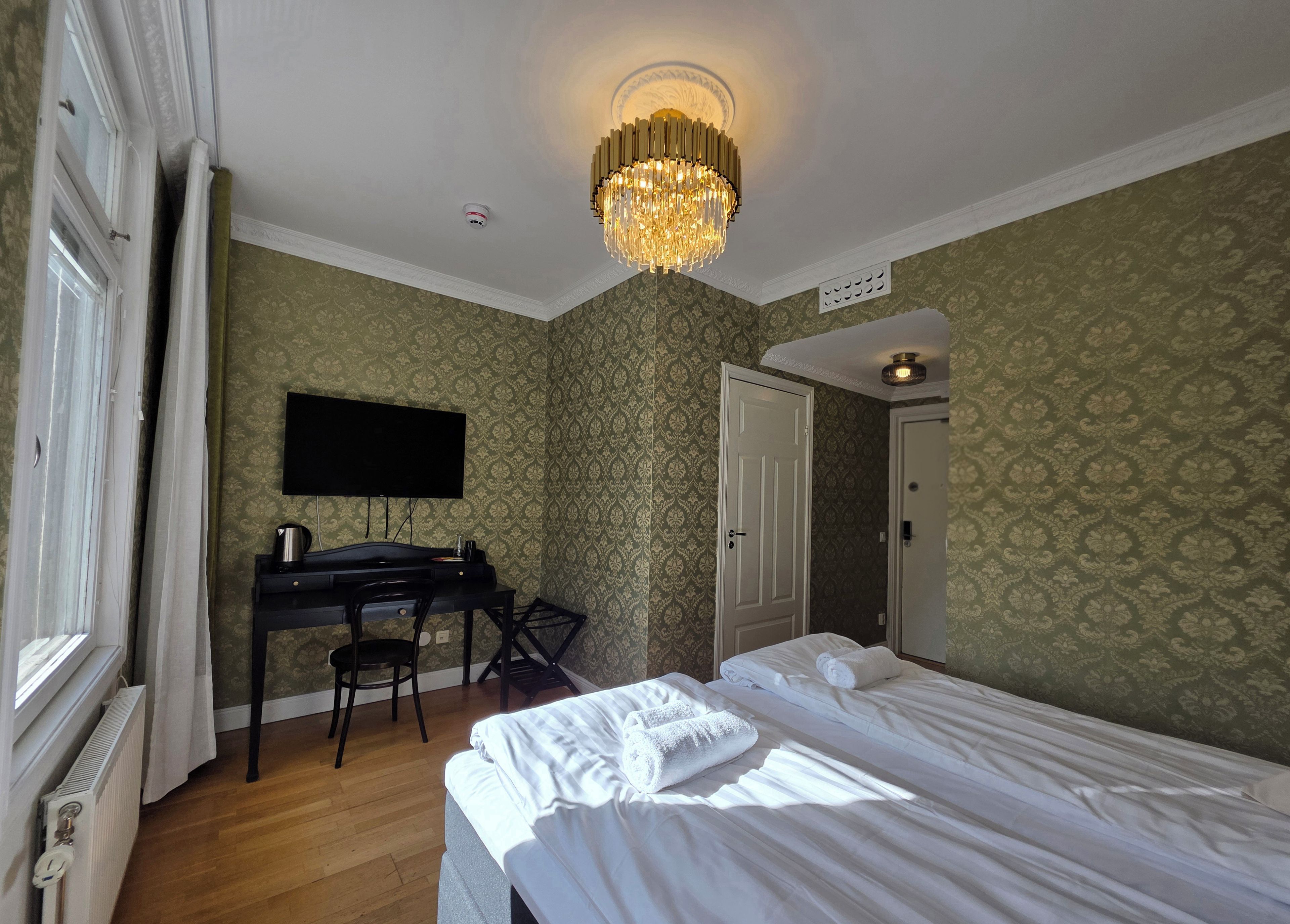 A hotel room with a bed, a desk with a flat-screen TV, a chandelier, and patterned wallpaper.