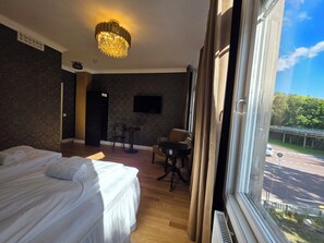 A hotel room with a bed, a desk, a chair, a television, and a view of a road and greenery outside.