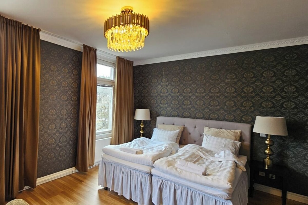A bedroom with two beds, a chandelier, and a view of trees through large windows.