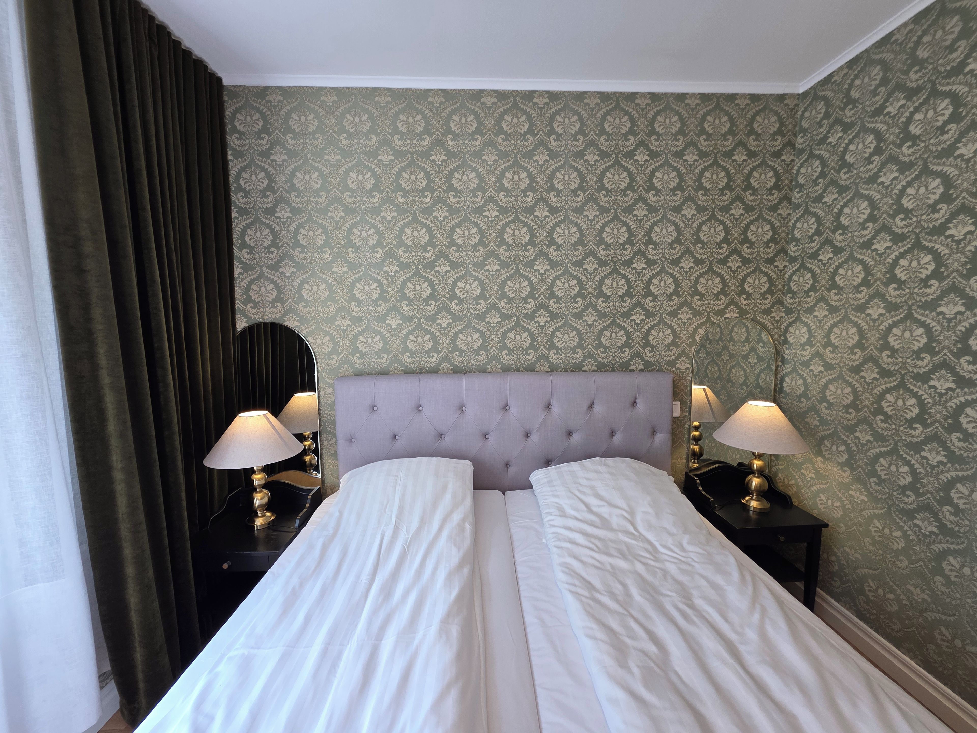 Una habitación de hotel con una cama, dos mesitas de noche con lámparas, un espejo y un papel tapiz estampado.