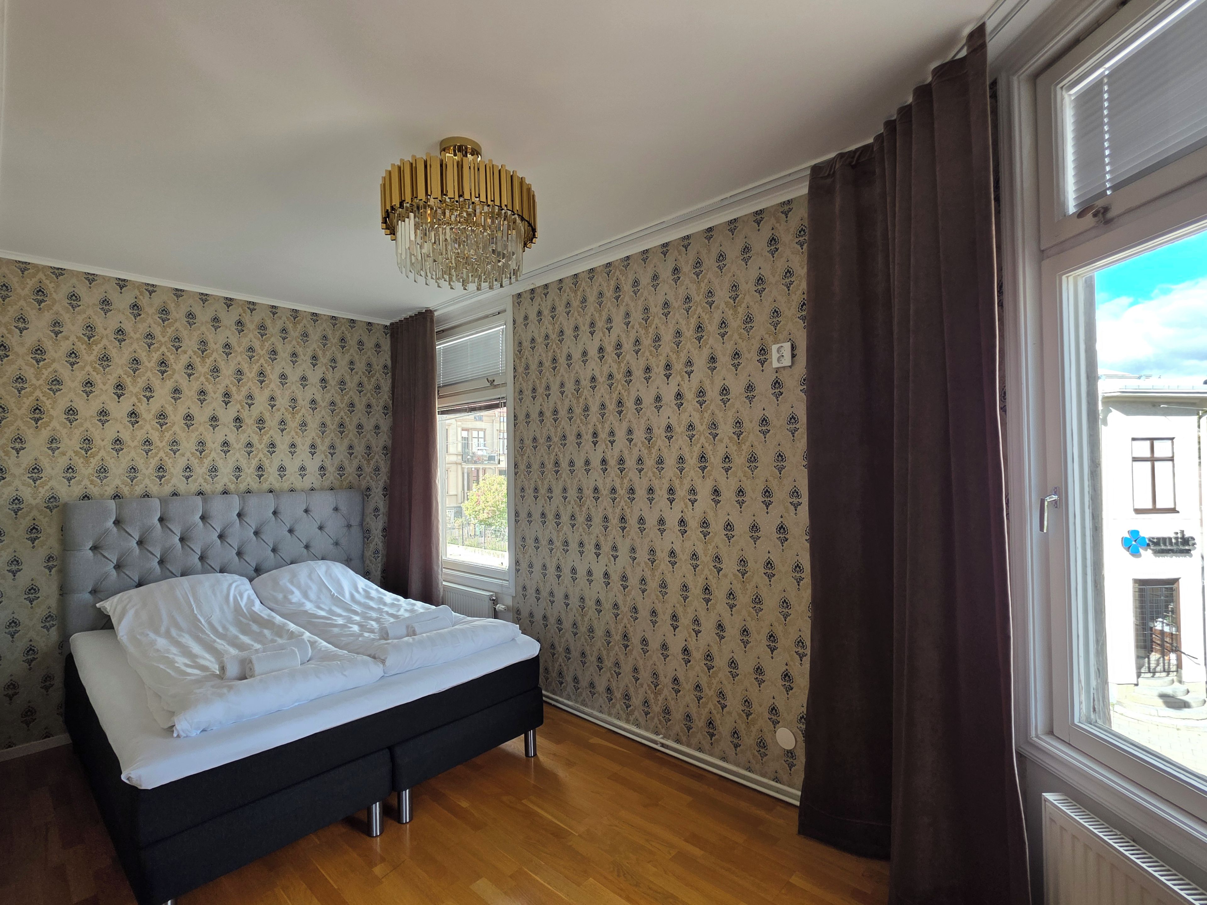 Un dormitorio con cama, candelabro, papel tapiz estampado y ventana con cortinas.