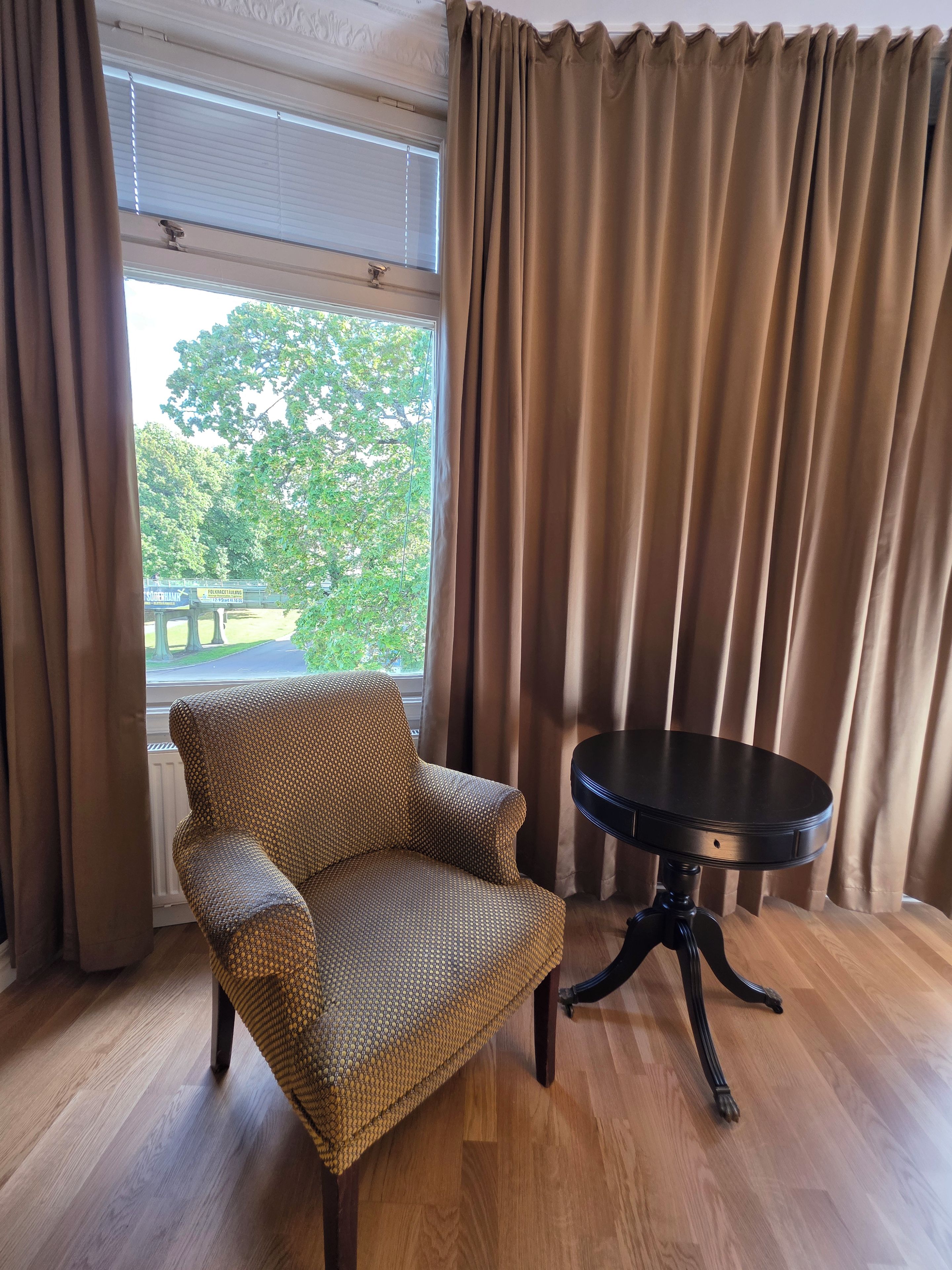 A room with a patterned armchair, a round table, and a view of a tree outside through a window with curtains.