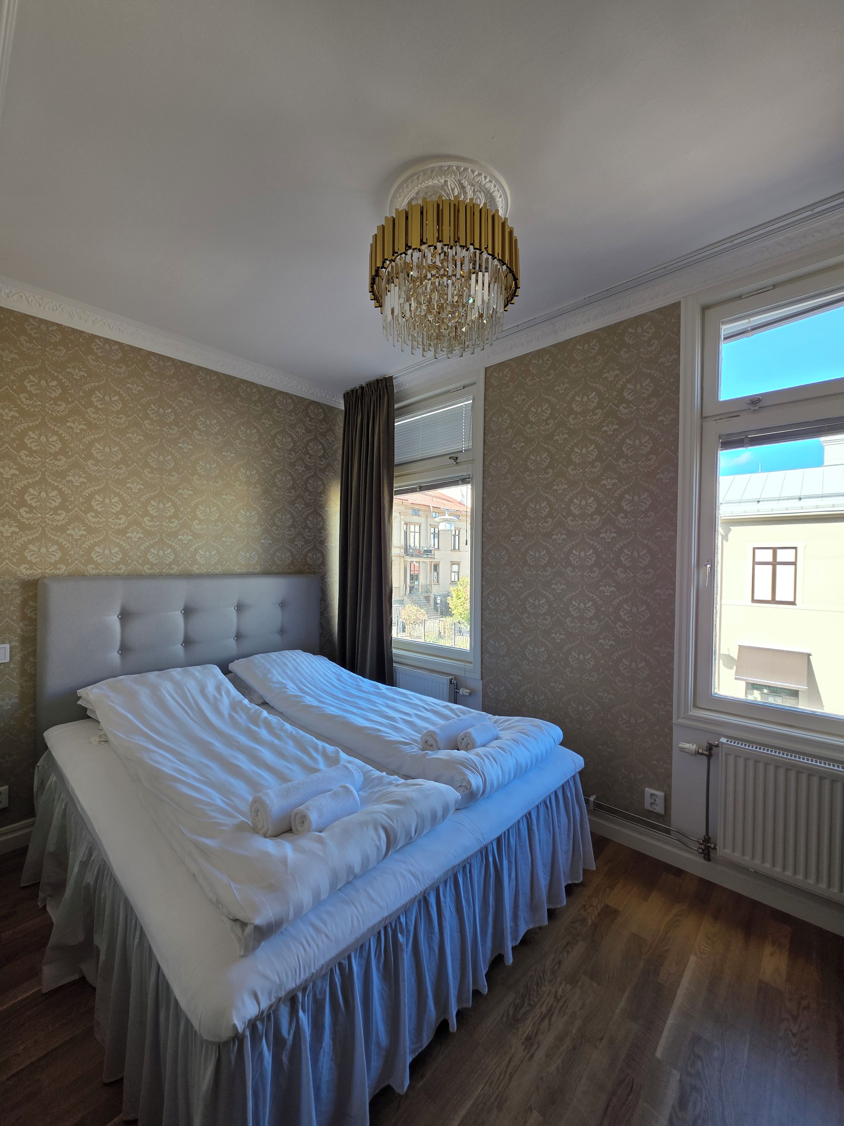 Habitación de hotel con una cama grande, un candelabro, un ventanal con vistas a edificios y papel tapiz estampado.