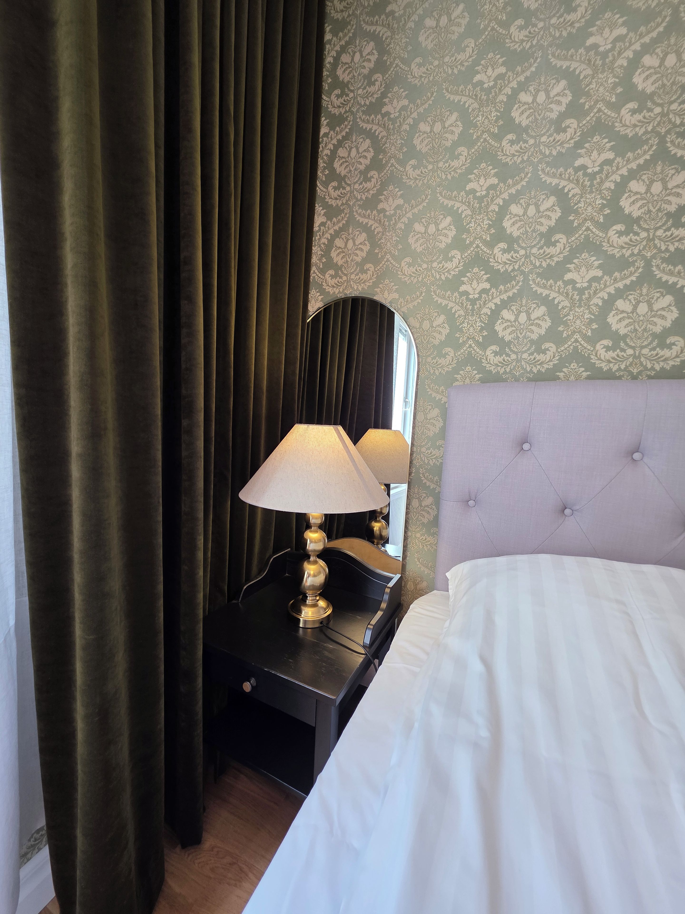 A hotel room with a bed, a nightstand with a lamp, a mirror, and a window with curtains.