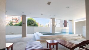 A modern outdoor lounge area with wooden loungers, a small pool, and a view of the cityscape. - Acqua Prime (Maceió)