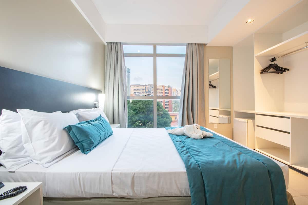 Quarto de hotel com uma cama grande, um guarda-roupa, um espelho e janela com vista para a cidade.