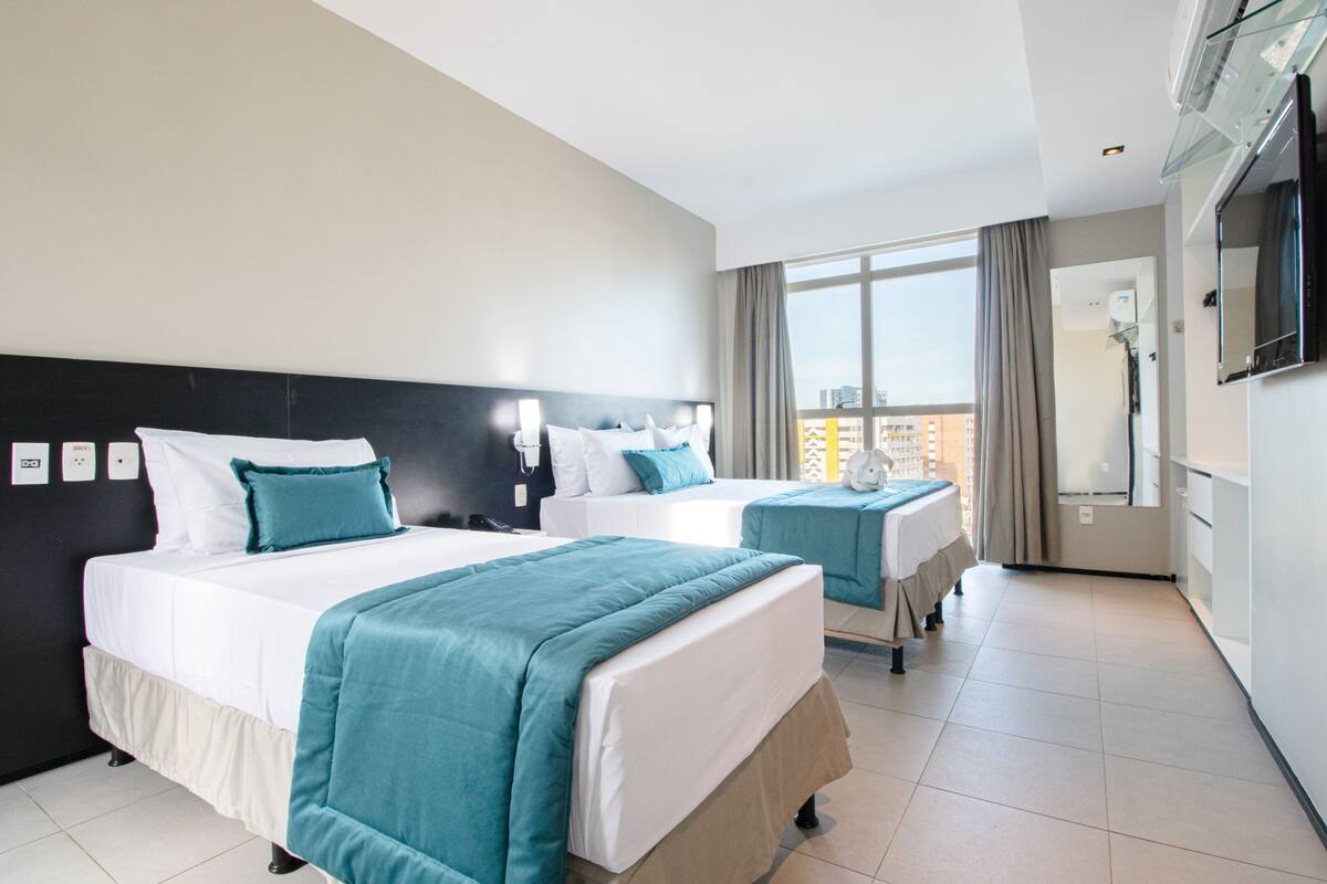 Quarto de hotel com duas camas, uma janela grande e uma televisão na parede.