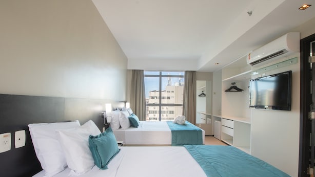 A hotel room with two beds, a TV, a wardrobe, and a window with a city view. - Acqua Prime (Maceió)