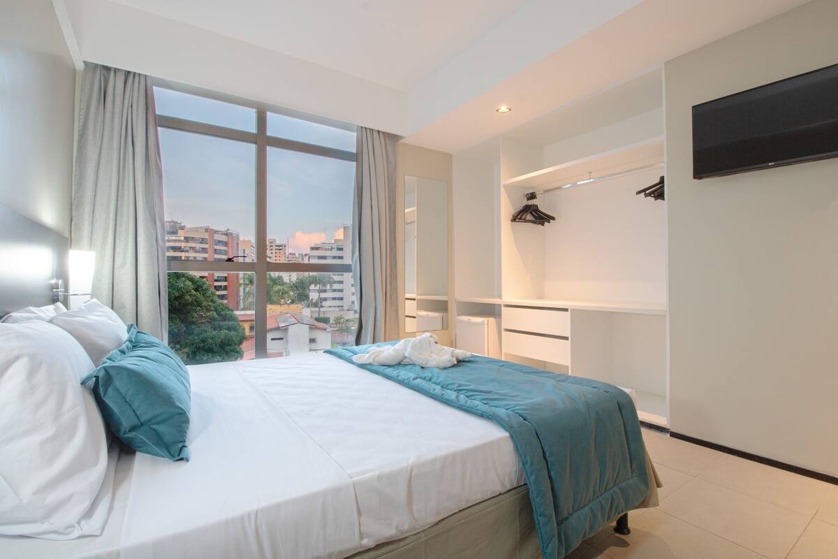 Quarto de hotel moderno com uma cama grande, vista para a cidade, televisão na parede e varanda com grade.