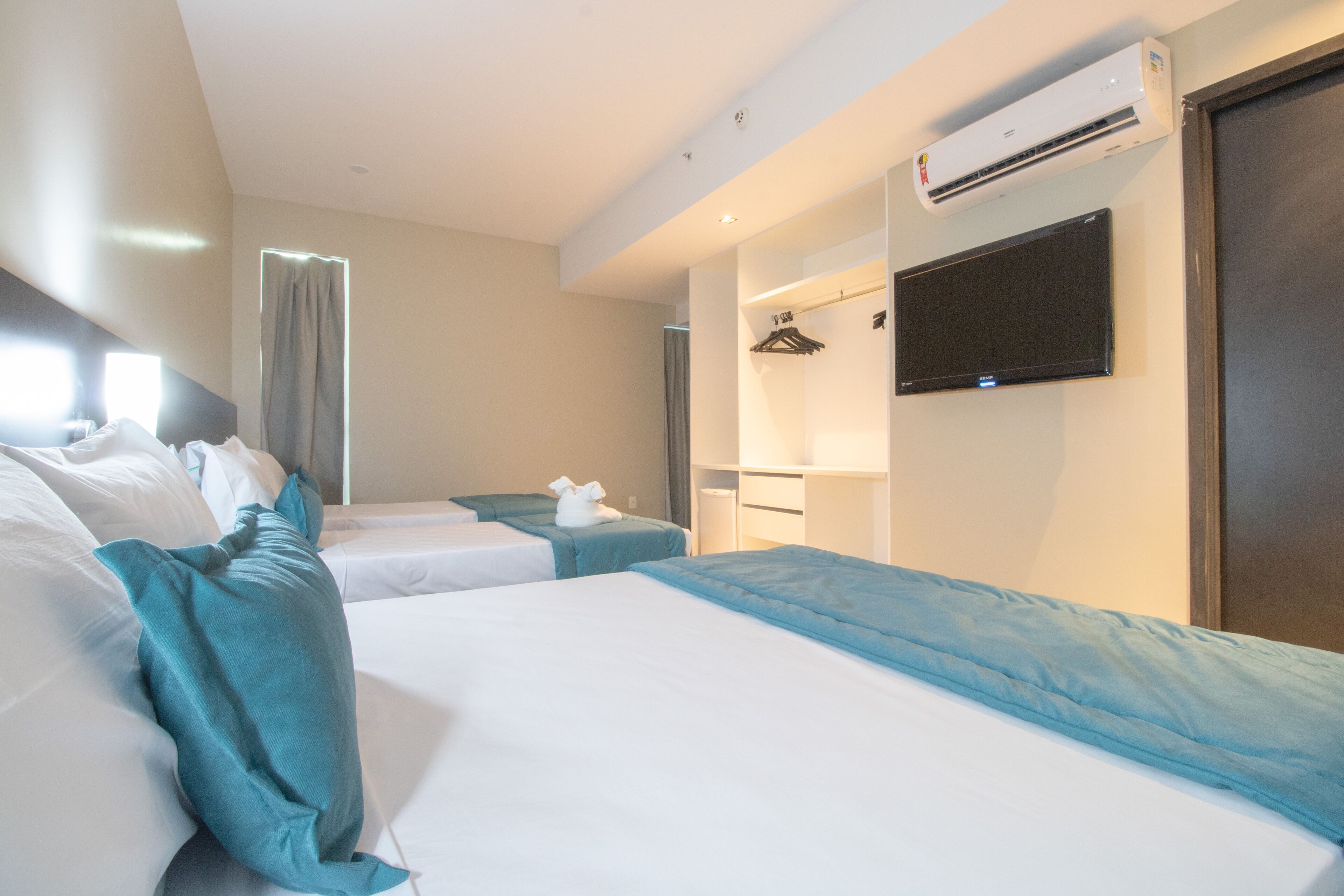 A hotel room with two beds, a flat-screen TV, an air conditioning unit, and a window with curtains.