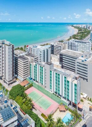 Um complexo de edifícios altos com quadra de tênis e piscina, com vista para uma praia e oceano.