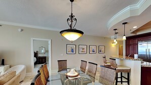 Zona pranzo con tavolo in vetro, sedie in vimini, un lampadario e quadri appesi al muro.