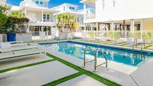 Una piscina con una escalera de metal, rodeada por un suelo de hormigón con bordes de césped, sillas de descanso blancas y una valla blanca.