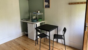 A compact kitchen with a white refrigerator, a small sink, a black table, and two black chairs. A white shelf unit with dishes and a bowl is mounted on the wall. - Etretat Aventure (Les Loges)