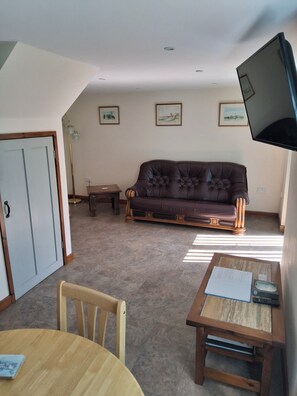 Una sala de estar con un sofá de cuero marrón, una mesa de comedor de madera, un televisor montado en la pared y una puerta.