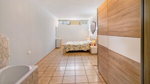 A narrow, tiled bathroom with a bathtub, a bed with a floral bedspread, and a wooden panel wall.