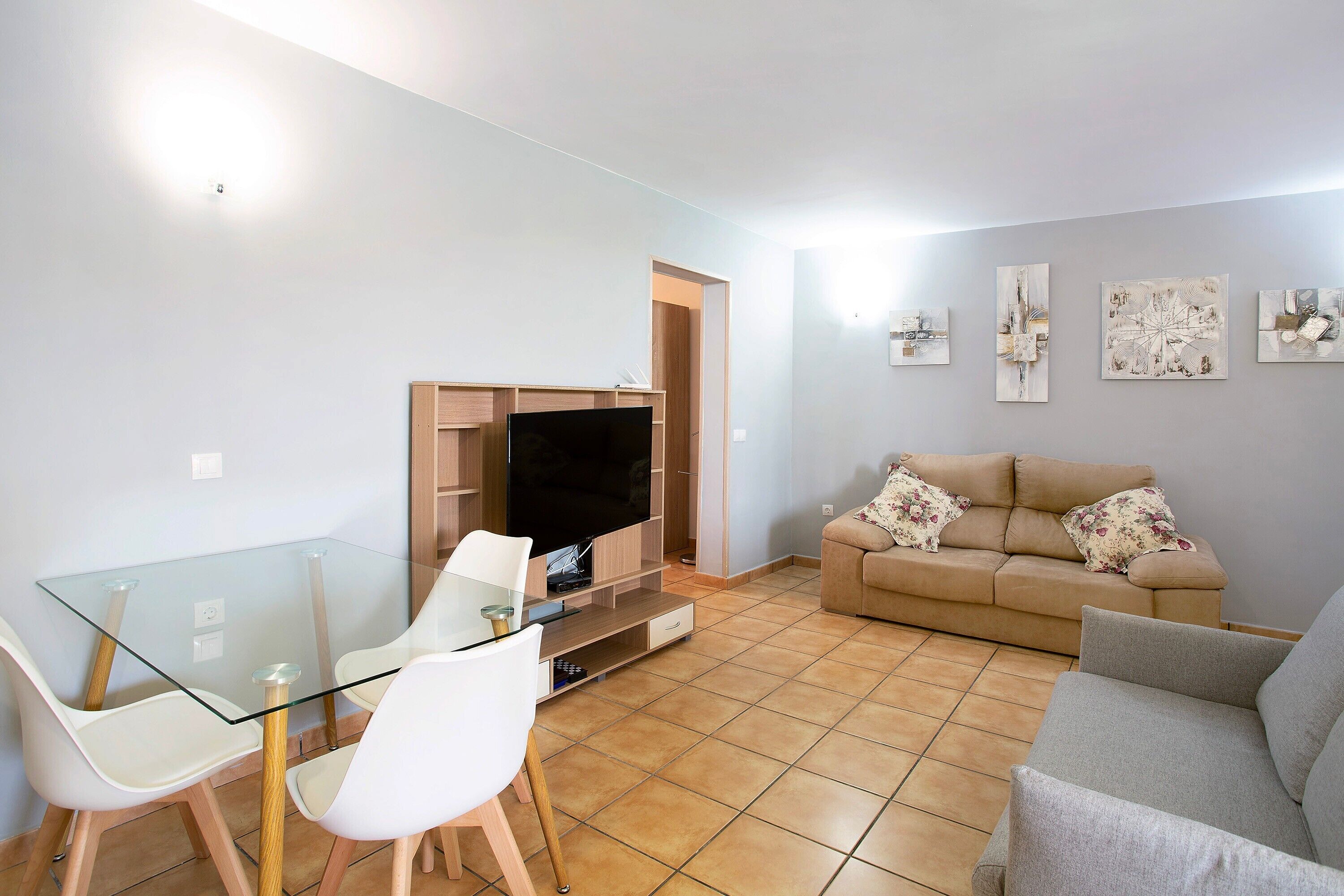 Un salon avec une table en verre, deux chaises blanches, un canapé beige, un téléviseur à écran plat et des œuvres d’art accrochées au mur.