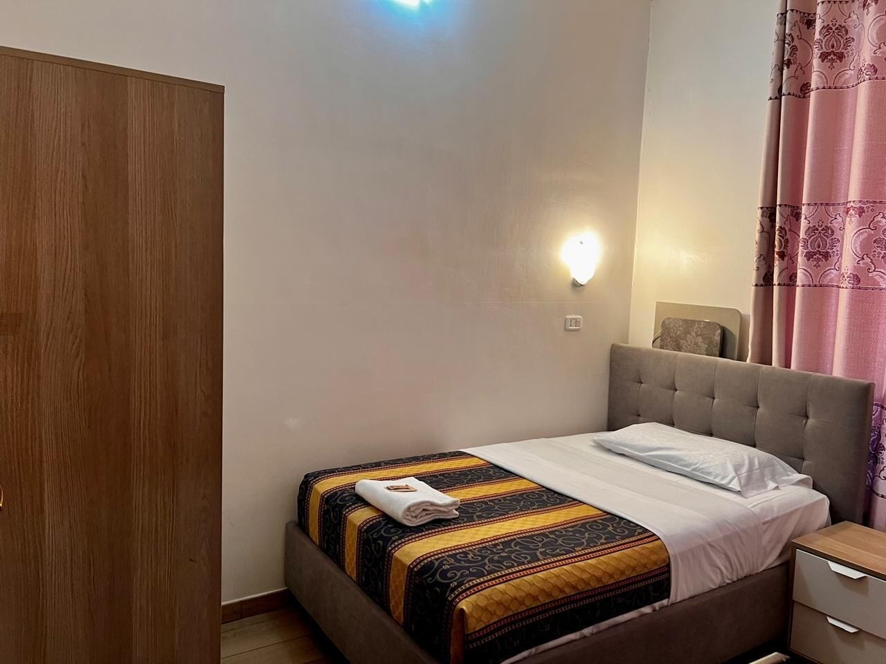 A single bed with a patterned bedspread, a bedside table, a nightstand, a wall-mounted light, and a wooden wardrobe.