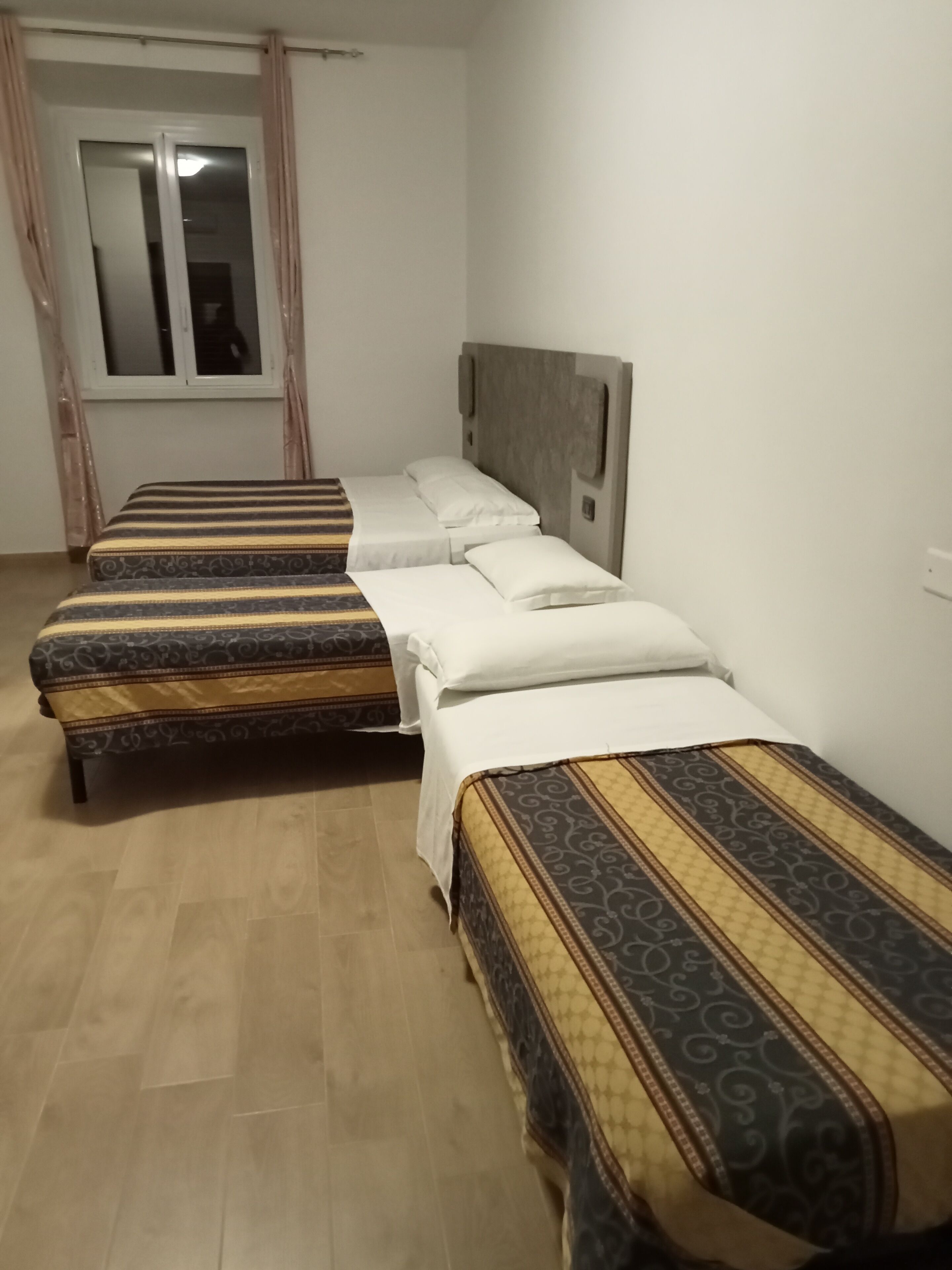 Habitación de hotel con tres camas individuales, cada una con ropa de cama estampada, suelo de madera, ventana con cortinas y pared blanca lisa.