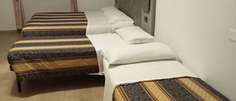 Habitación de hotel con tres camas individuales, cada una con ropa de cama estampada, suelo de madera, ventana con cortinas y pared blanca lisa.