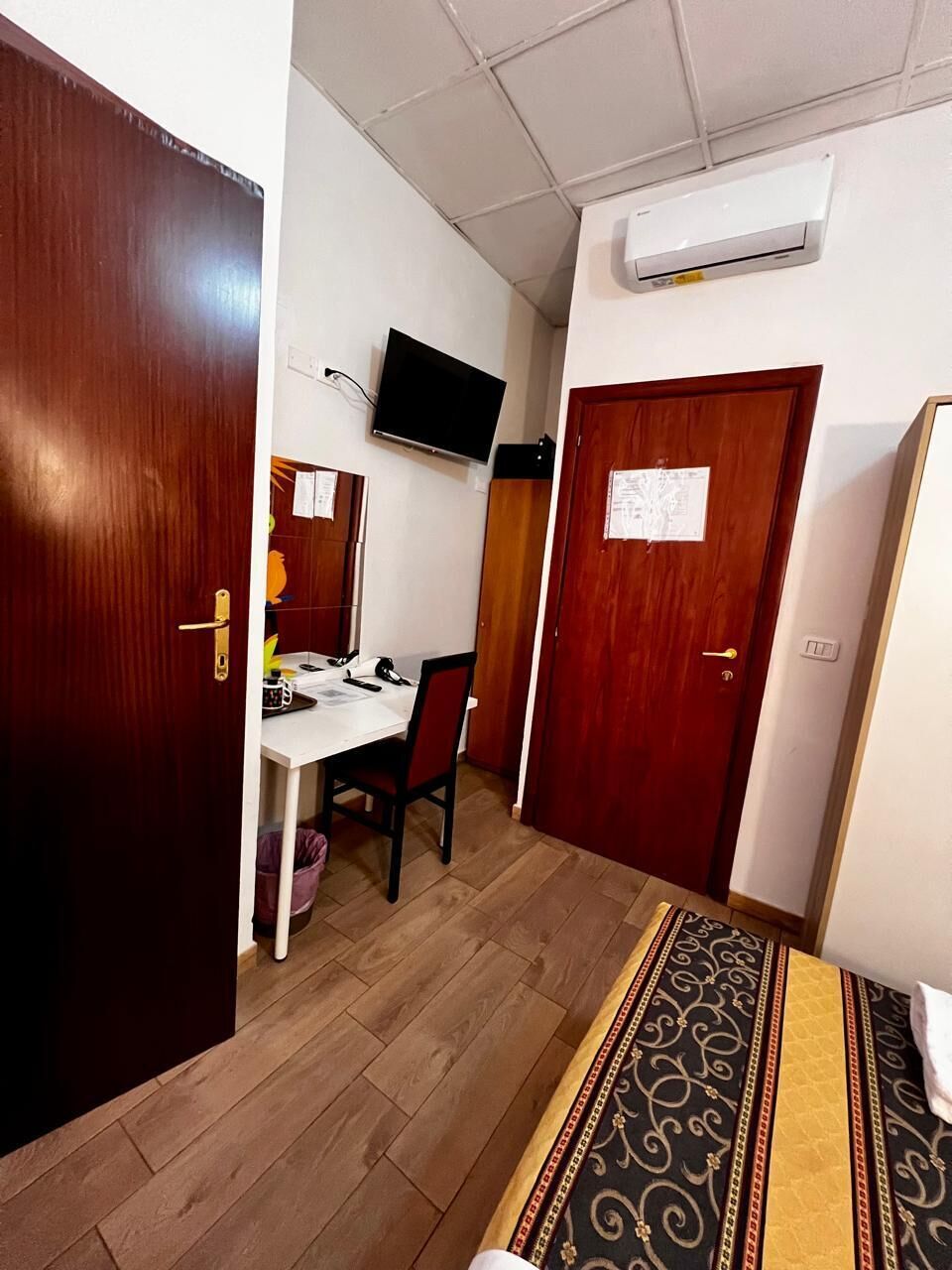 A room with a wooden door, a desk with a chair, a television on the wall, and an air conditioning unit.