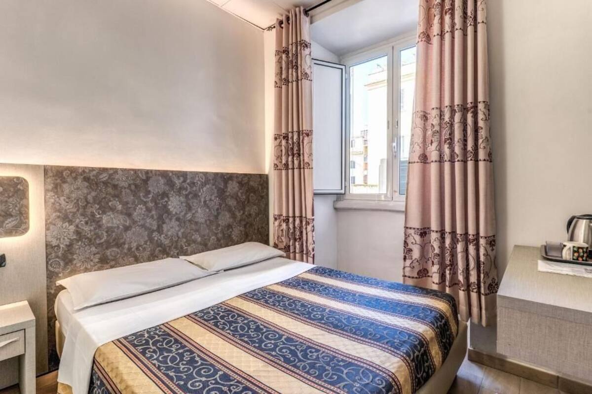 A hotel room with a bed, a nightstand, a window with curtains, and a view of buildings outside.