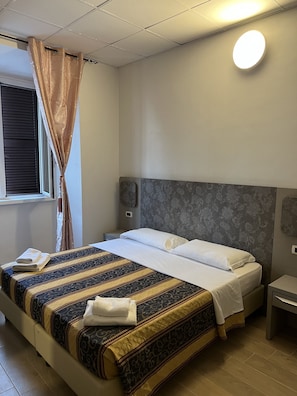A neatly made bed with a patterned bedspread, a bedside table, a wall-mounted light, and a window with curtains. - HOTELPRINCIPE (Rome)