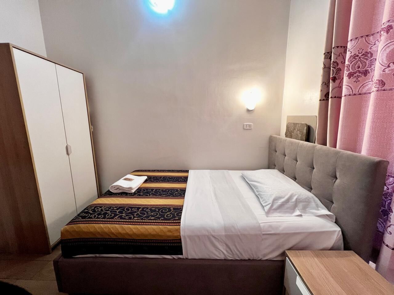 A single bed with a headboard, a bedside table, a wardrobe, and a wall-mounted light fixture.