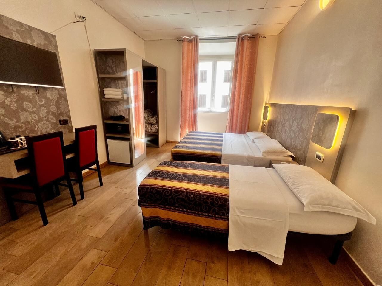 A hotel room with two beds, a desk with chairs, a wardrobe, and a window with curtains.
