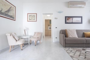 A living room with a sofa, two armchairs, a small table, and a rug. There are framed pictures on the walls, a doorway leading to a kitchen, and an air conditioning unit on the wall.