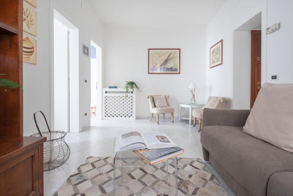 A living room with a sofa, a coffee table with a book, a chair, and a small table with a lamp. There is a plant on a white console table and a decorative wall hanging.