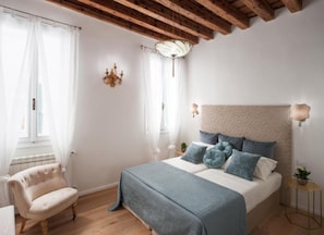 A bedroom with a bed, a chair, a chandelier, and a window with curtains.