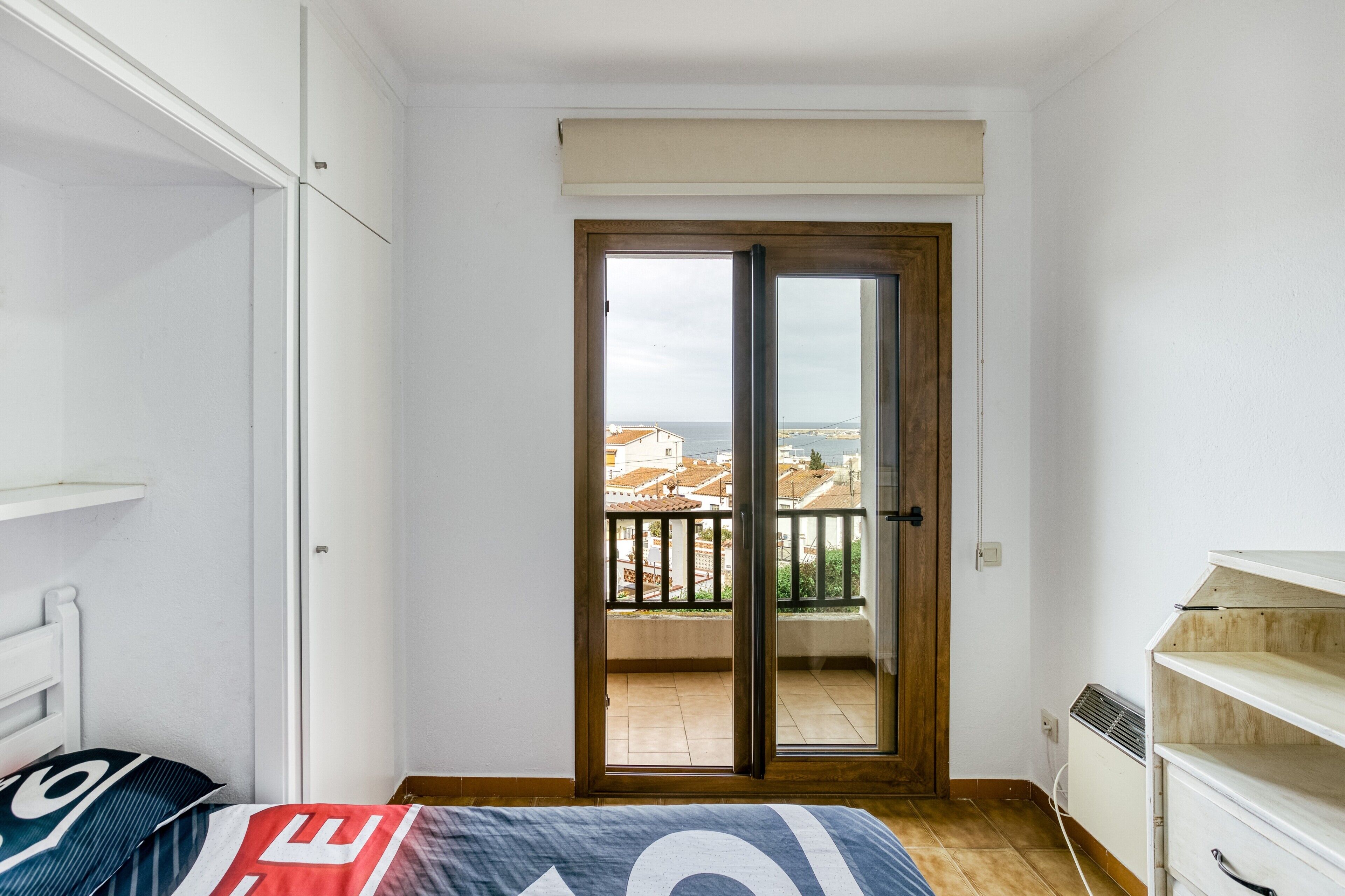 Un dormitorio con cama, armario, radiador y balcón con vistas a edificios.
