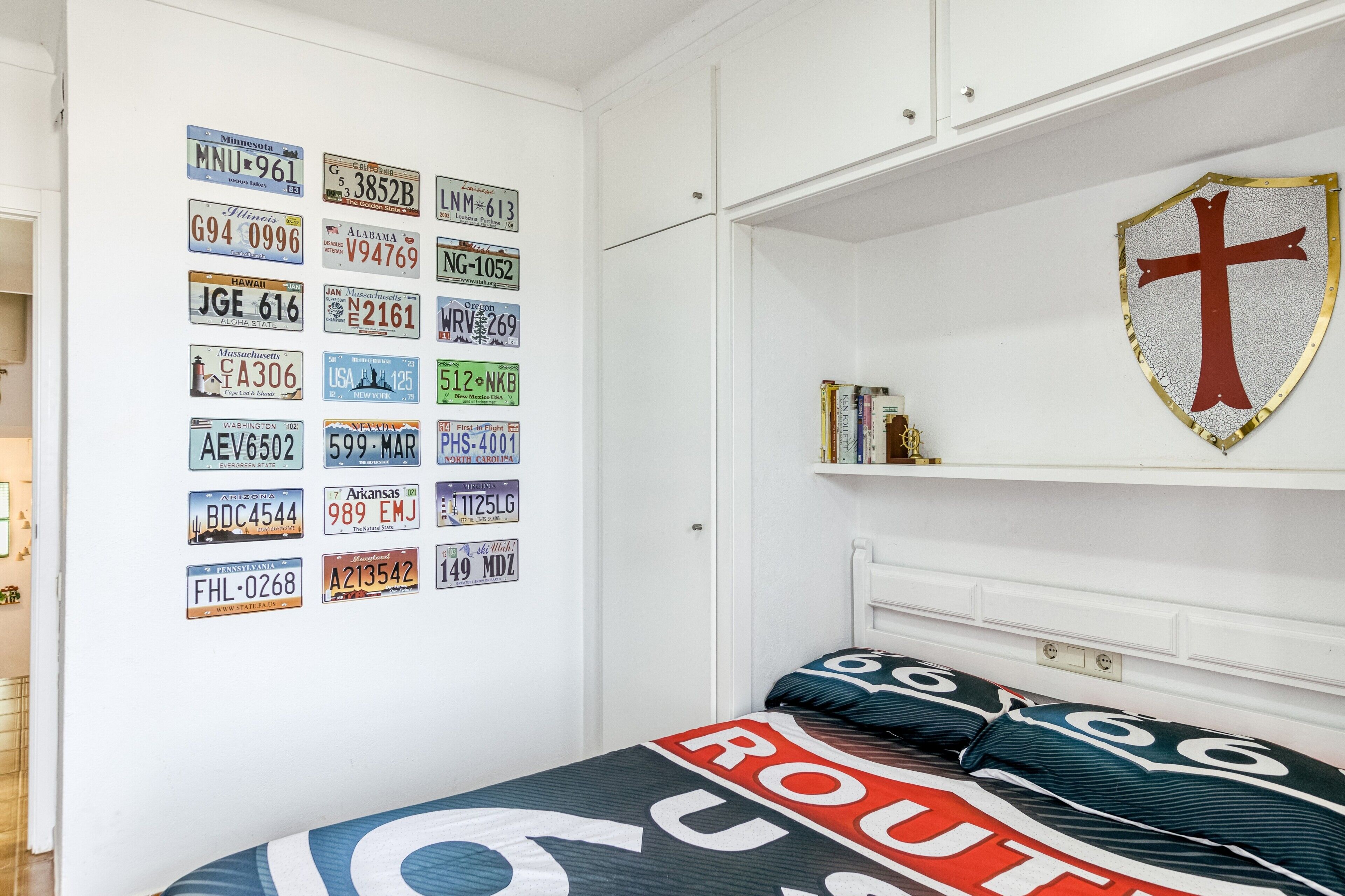 A bedroom with a bed, a wall of various state license plates, a cross shield decoration, and a shelf with books.