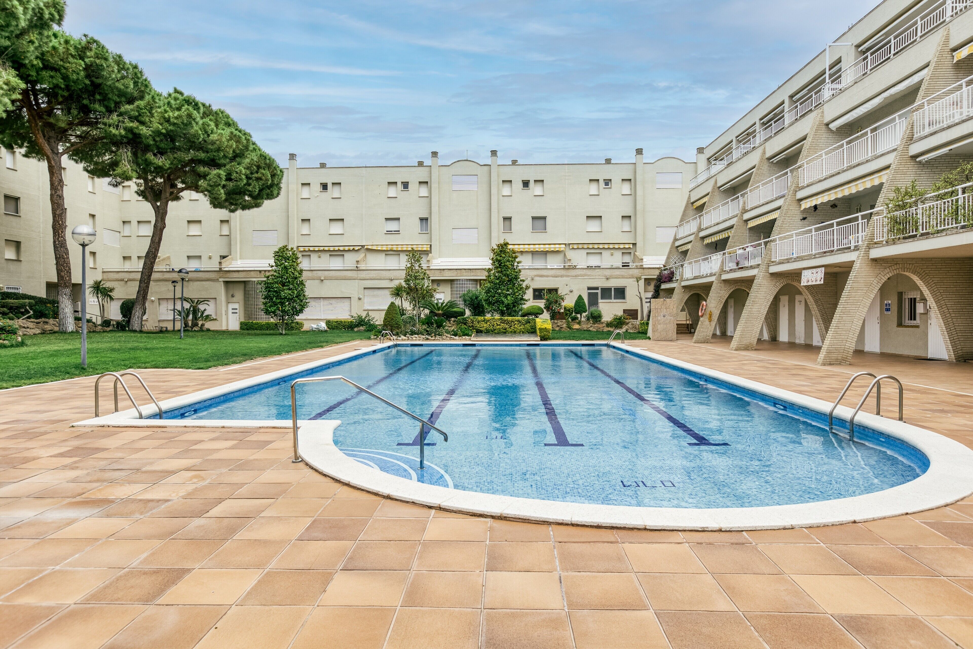 Una piscina con plataforma empedrada, rodeada por un edificio con varios balcones y un jardín.