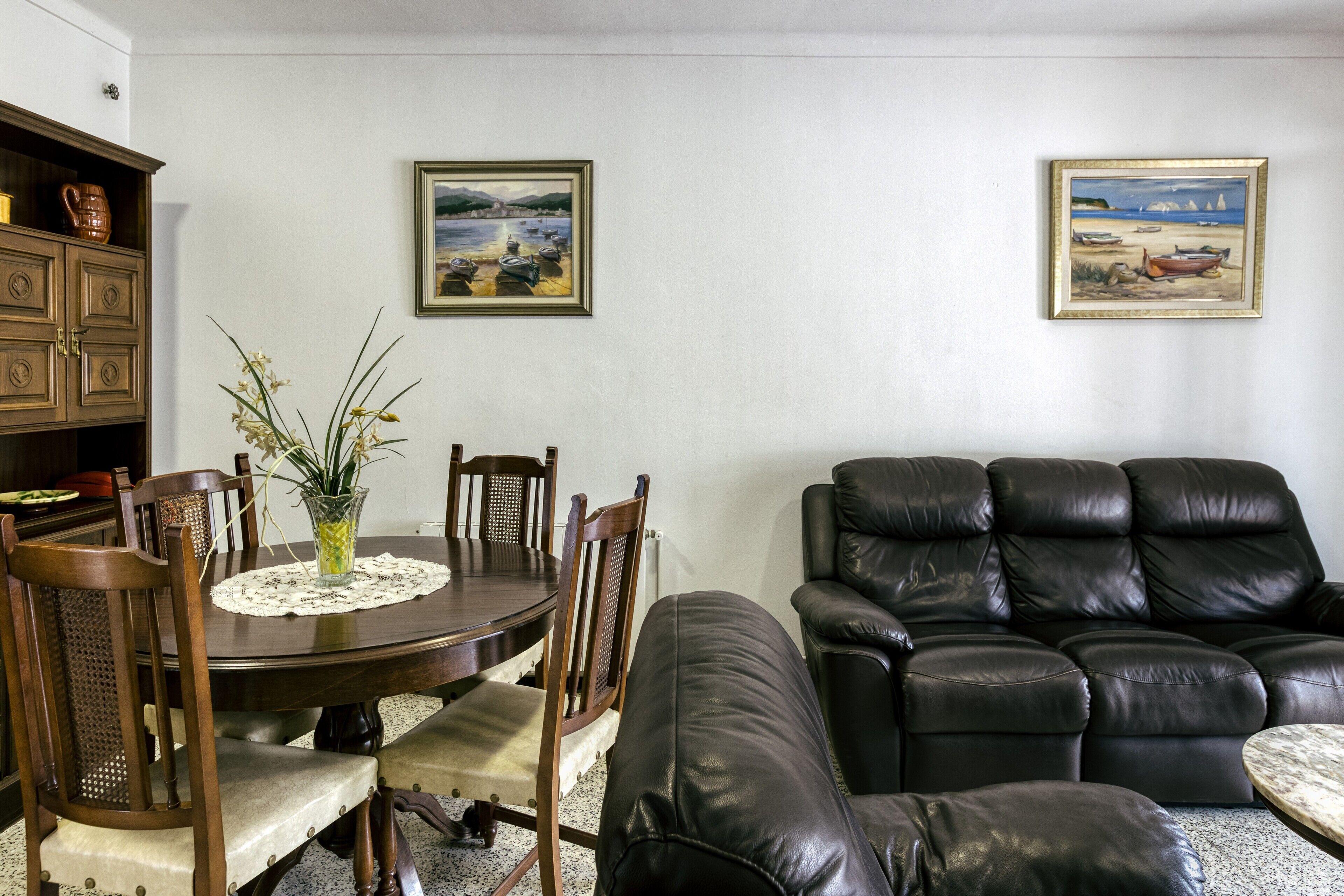 A dining area with a round table, chairs, and a vase of flowers. There is a black leather sofa and a cabinet with dishes. Two framed paintings are on the wall.