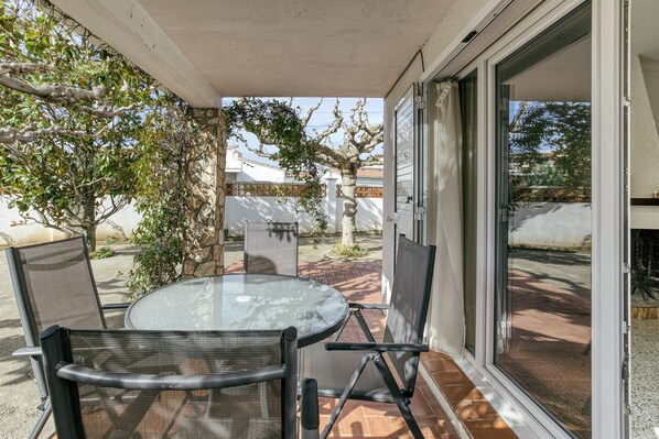 A patio with a glass table and chairs, a sliding glass door, and a view of a tree and fence.