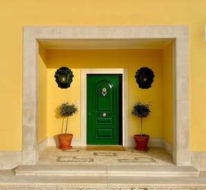 Un edificio amarillo con puerta verde, dos macetas y dos luces fijadas a la pared.