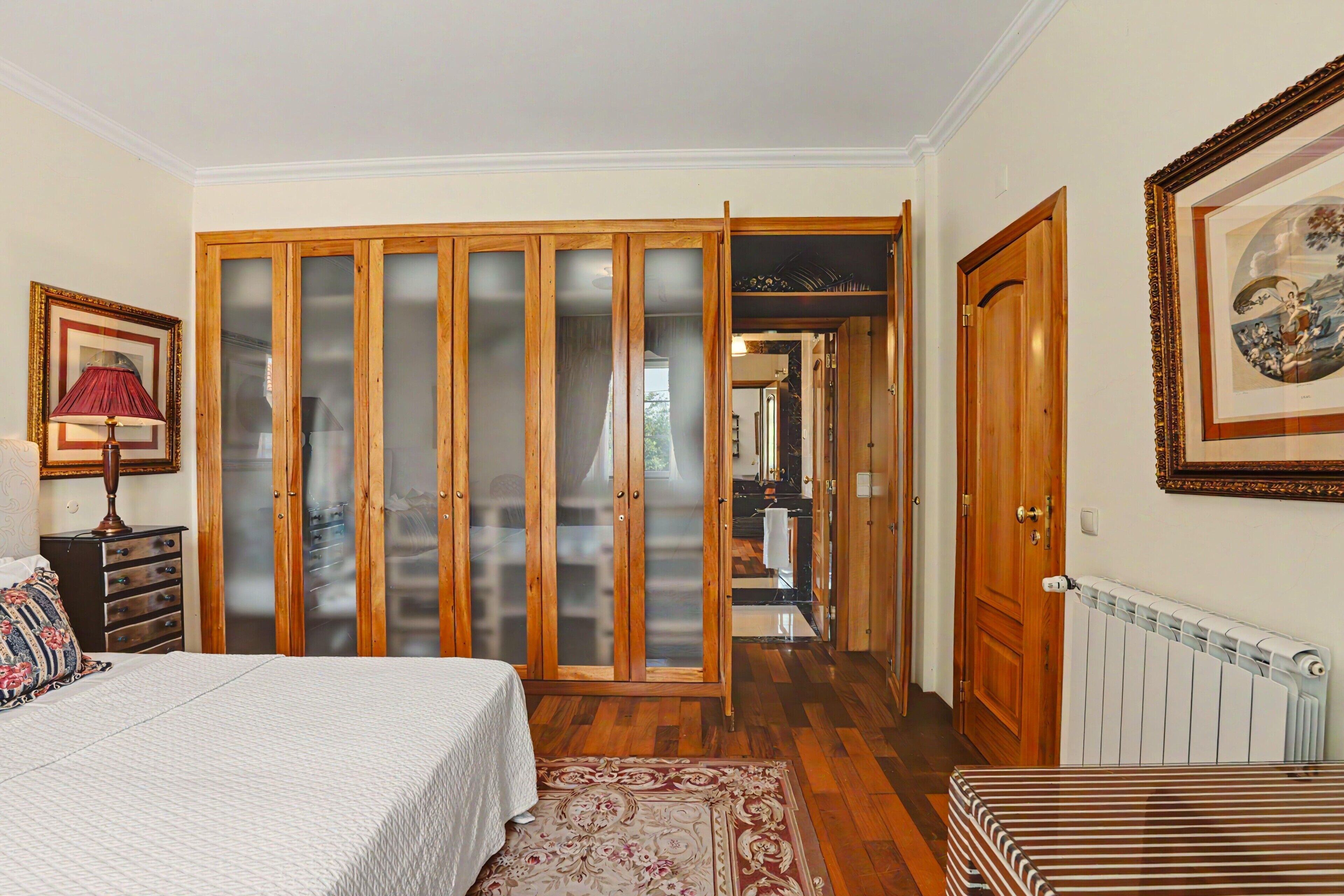 A bedroom with a bed, a dresser, a nightstand, a lamp, a wardrobe, and a radiator.