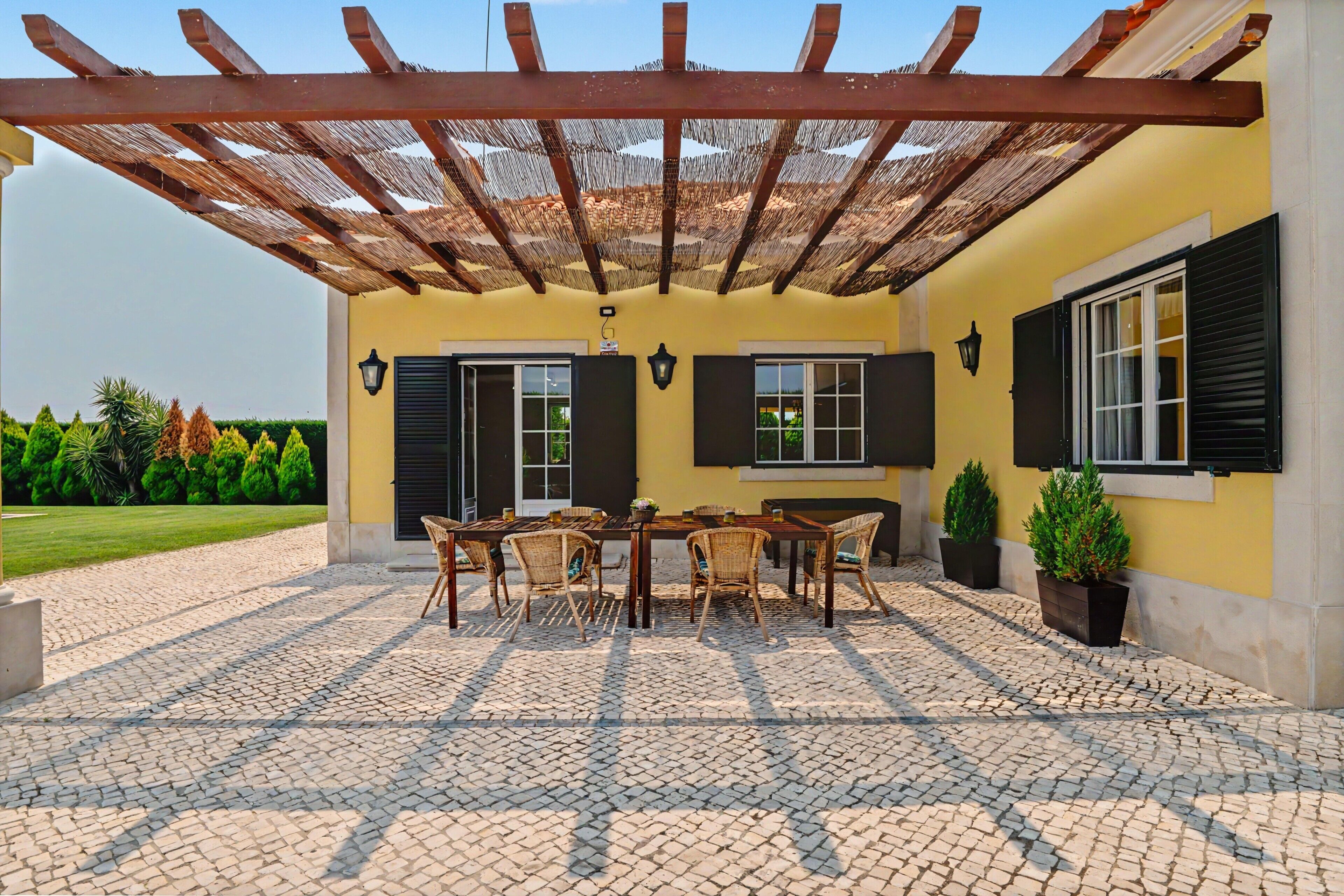 Un patio exterior techado con mesa y sillas, una pérgola y plantas en macetas.
